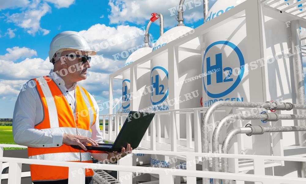 На фотографии изображен инженер в защитной каске и оранжевом жилете, работающий с ноутбуком на фоне водородных резервуаров на заправочной станции. Это изображение подчеркивает важность мониторинга и управления в процессе работы водородных заправочных станций, иллюстрируя современные технологии в области возобновляемой энергетики.